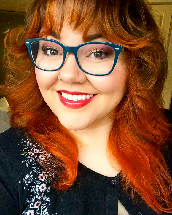 Machine embroidery help for beginners Picture of Kate, a woman with long wavy neon orange hair and bangs. She has teal glasses, a big smile and a black cardigan embroidered with white flowers.